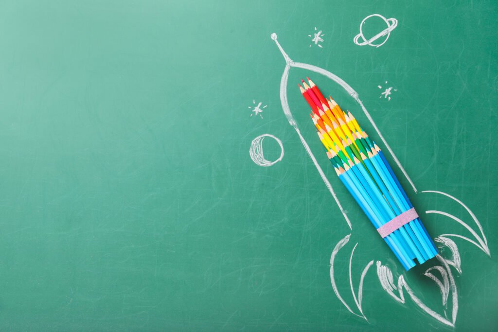 A space rocket drawn in white chalk on a green board with coloured pencils filling the body of the rocket